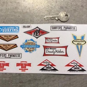 May include: A sheet of vintage-style surf stickers with various designs, including "Surfers Paradise", "I'd rather be surfing", "Bear", "Dewey Weber", and "Greek".