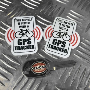 Puede incluir: Dos pegatinas blancas con texto negro y un gráfico de bicicleta roja. El texto dice "Esta bicicleta está equipada con un rastreador GPS". Las pegatinas están sobre una superficie de metal plateado. Una llave negra con una pegatina negra y roja que dice "Oil Cat Stickers" está en primer plano.