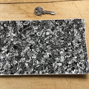 Op de afbeelding: Een rechthoekig vel bedekt met een zwart-wit stickerpatroon, met diverse afbeeldingen en tekst, waaronder "BOOST" en "DRIFTING." Een zilveren sleutel ligt boven het vel op een houten oppervlak. Het sticker vel is waarschijnlijk voor auto- of decoratief gebruik.