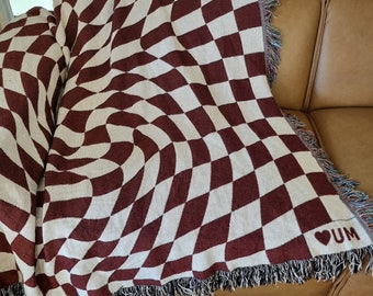 Puede incluir: Una manta tejida de color granate y crema con un patrón de tablero de ajedrez deformado. La manta tiene un borde con flecos y las letras "UM" con un símbolo de corazón son visibles en la esquina. La manta está sobre un sofá de cuero marrón.
