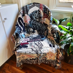 Puede incluir: Una manta de tapiz con un dise&ntilde;o de tigre blanco y negro, con un fondo blanco con acentos negros y rojos. La manta tiene un borde de flecos y est&aacute; drapeada sobre una silla.