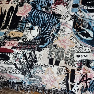 Puede incluir: Una manta de tejido de tapicer&iacute;a con un esquema de color negro, blanco, azul y rosa. La manta presenta un dise&ntilde;o gr&aacute;fico de un tigre, flores y texto. El texto incluye las palabras "are" y "UM".