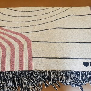 May include: A cream-colored woven throw blanket with black and pink stripes and a black heart with the text "UM" on the bottom right corner. The blanket has black fringe around the edges.