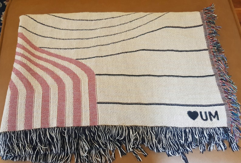 May include: A cream-colored woven throw blanket with black and pink stripes and a black heart with the text "UM" on the bottom right corner. The blanket has black fringe around the edges.