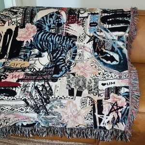 Puede incluir: Una manta de tejido de tapicer&iacute;a con un dise&ntilde;o abstracto en negro, blanco y azul que presenta un tigre, flores y las palabras "are my" y "UM".