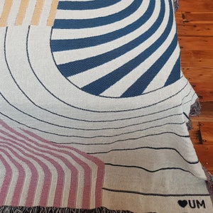 May include: A woven throw blanket with a geometric pattern in shades of blue, pink, yellow, and white. The blanket has a fringe border and a small heart with the word "UM" in black.
