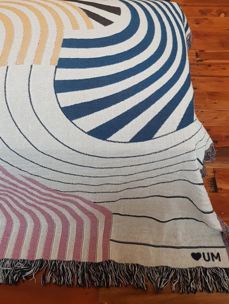 May include: A woven throw blanket with a geometric pattern in shades of blue, pink, yellow, and white. The blanket has a fringe border and a small heart with the word "UM" in black.