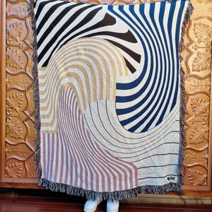 May include: A woven tapestry throw blanket with a geometric pattern in black, white, blue, gold, and pink. The blanket has a fringe border and the word "RUM" is woven into the bottom right corner.