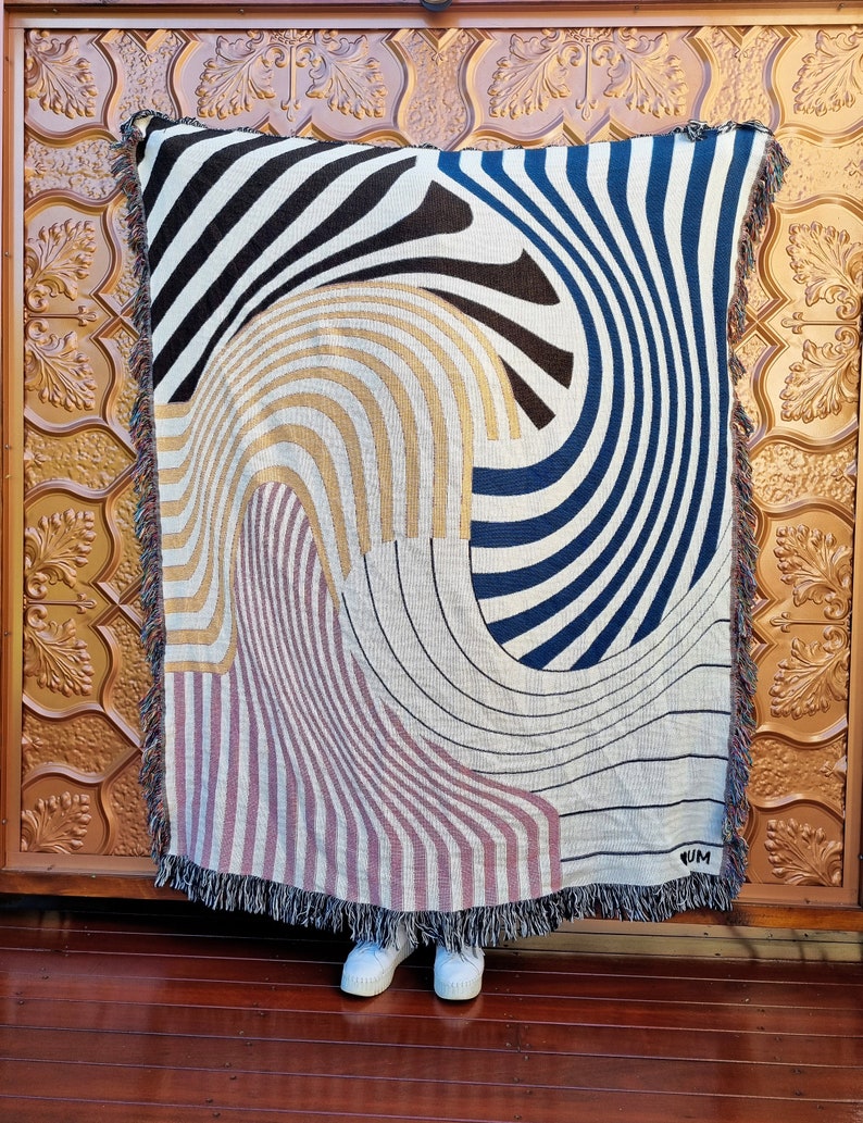 May include: A woven tapestry throw blanket with a geometric pattern in black, white, blue, gold, and pink. The blanket has a fringe border and the word "RUM" is woven into the bottom right corner.
