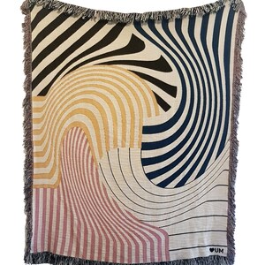 May include: A woven throw blanket with a geometric pattern in shades of brown, black, yellow, pink, and white. The blanket has a fringe border and the text "&hearts; UM" in the bottom right corner.