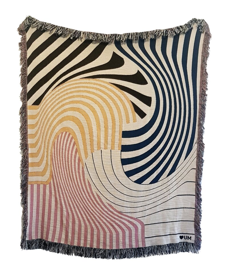 May include: A woven throw blanket with a geometric pattern in shades of brown, black, yellow, pink, and white. The blanket has a fringe border and the text "&hearts; UM" in the bottom right corner.