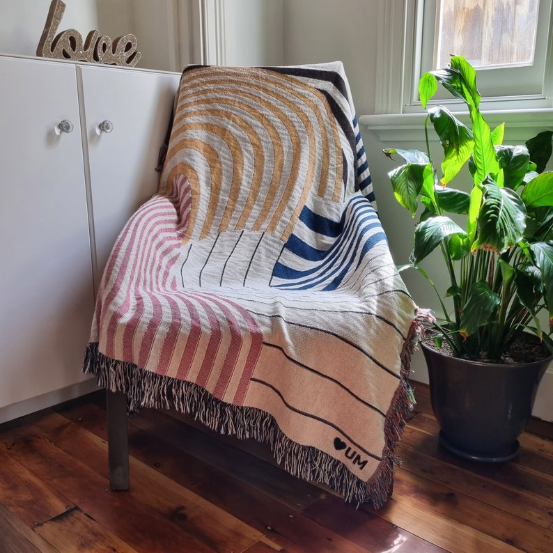 May include: A multi-colored throw blanket with a fringe border. The blanket is draped over a chair and features a geometric pattern with stripes and curves in shades of pink, yellow, blue, and white. The blanket has a heart with the letters "UM" inside.