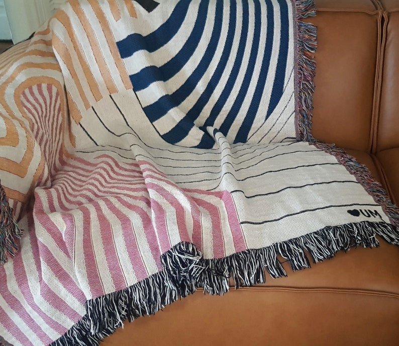 May include: A woven throw blanket with a geometric pattern in shades of orange, blue, pink, and white. The blanket has fringe on all sides and features the text "I &hearts; UM" in black.