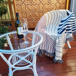 May include: A white wicker table with a glass top and a bottle of wine and a glass on the table. A striped throw blanket is draped over a white wicker chair.