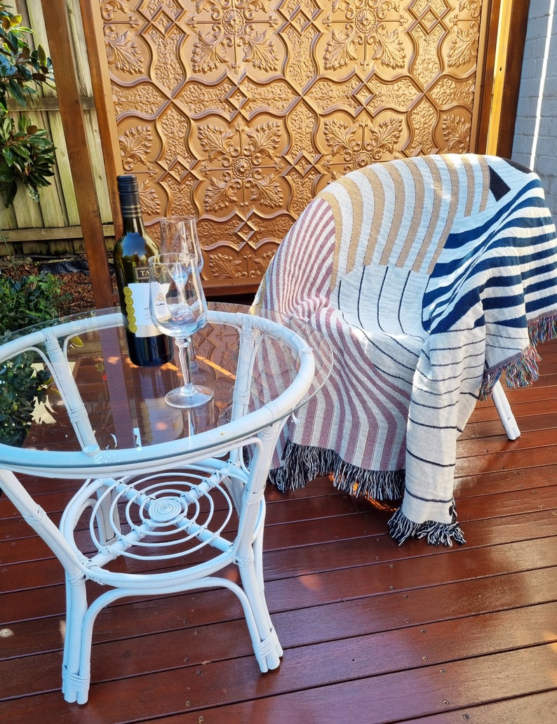 May include: A white wicker table with a glass top and a bottle of wine and a glass on the table. A striped throw blanket is draped over a white wicker chair.