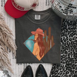 May include: A dark gray t-shirt featuring a desert-themed graphic of a woman in a cowboy hat and cacti. A red and tan baseball cap with the word "TIMELESS" is visible. Black cowboy boots and a leopard print fabric are also in the image.