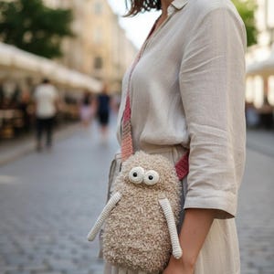 May include: A beige, handmade crossbody bag shaped like a whimsical monster. The bag has large, round eyes, long arms, and legs. The strap is pink and beige. The bag is made of a textured, fuzzy material.