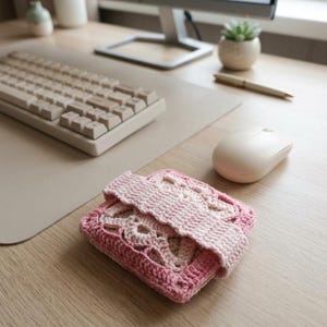 May include: A handmade, pink crochet case with an openwork design, resting on a wooden desk. The case is square with a folded flap. A computer keyboard, mouse, and monitor are in the background.