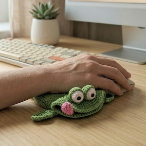 May include: A green, crocheted frog mouse mat with large, round eyes and a pink nose. The frog is on a wooden desk, with a computer mouse resting on it. A hand is using the mouse.