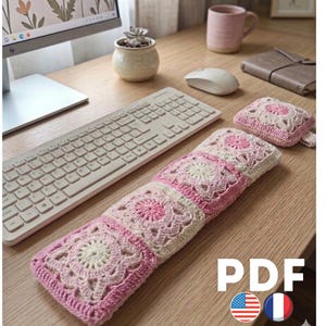 May include: A handmade crochet keyboard wrist rest and matching mouse pad in shades of pink and cream. The square pattern features a floral design. The image also shows a computer keyboard, mouse, and other desk accessories.