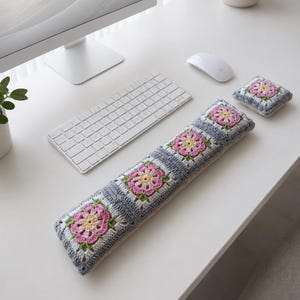 May include: A crocheted keyboard wrist rest and matching coaster. The items feature a floral design with pink, yellow, and green accents on a gray and white background. The wrist rest is rectangular and long, while the coaster is square.
