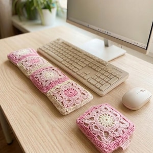 May include: A crocheted keyboard wrist rest and coaster set in pink and cream. The square designs have a floral pattern. The items are on a light wood desk next to a keyboard and mouse. The set is handmade.