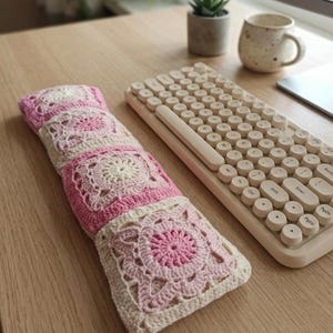 May include: A handmade crochet item in shades of pink and cream, featuring a floral design. The item is rectangular and appears to be a keyboard wrist rest. A cream-colored keyboard is next to the wrist rest on a wooden desk.