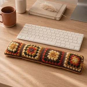 May include: A rectangular wrist rest for a computer keyboard, featuring a series of four joined crochet granny squares in a color palette of mustard yellow, burnt orange, dark brown, and cream, with a small loop on the right side.