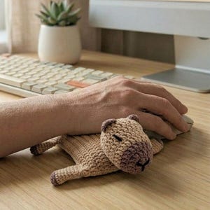 May include: A crocheted capybara-shaped mouse mat. The mat is mainly tan with a darker brown head and feet. The capybara has a small black stitched mouth and eyes. The mat is on a wooden desk.