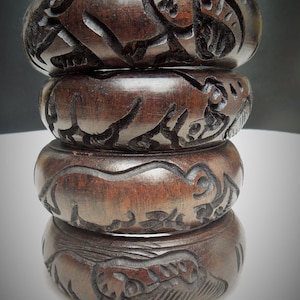 May include: Four dark brown wooden bangles with intricate carvings of animals, possibly elephants and rhinos. The bangles are stacked on top of each other.