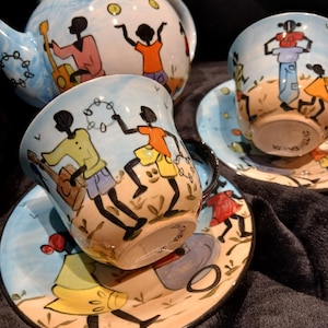 May include: A ceramic tea set featuring a teapot, two cups, and two saucers. Each piece is hand-painted with a light blue sky background and stylised black figures playing instruments, dancing, and juggling in colourful clothing on a tan ground.
