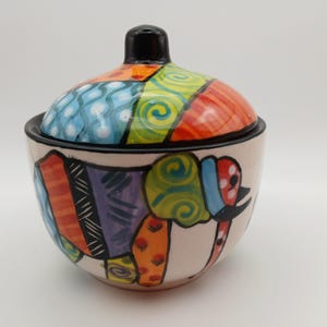 May include: A ceramic sugar bowl with a lid, featuring a hand-painted elephant design in vibrant colours including orange, purple, green, and blue. The bowl has a cream-coloured base and a black rim, with a matching lid topped by a black knob.
