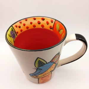 May include: A ceramic mug with a cream-coloured exterior featuring a colourful giraffe illustration. The interior is bright red, while the inner rim displays an orange band with small red dots. The handle is white with a black accent at the top.