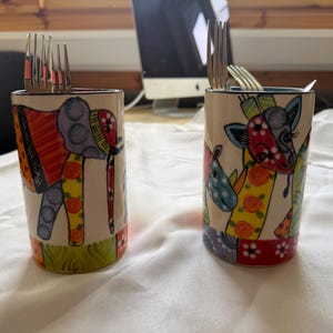 May include: Two cylindrical ceramic utensil holders featuring colourful, abstract animal illustrations. The left holder displays an elephant, while the right holder depicts a giraffe and rhinoceros. Both containers are filled with stainless steel forks.