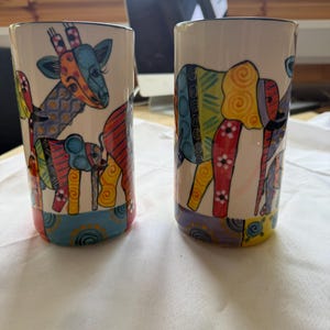 May include: Two cylindrical ceramic tumblers featuring colourful, abstract animal illustrations. The left tumbler displays a giraffe and a small elephant, while the right tumbler shows a large elephant. Both have vibrant, multi-coloured patterns.