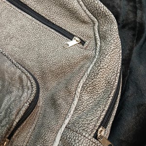 May include: Close-up of a grey, textured bag with black zips and silver zip pulls. The bag has a speckled pattern and a white seam. The background is a dark, textured fabric.