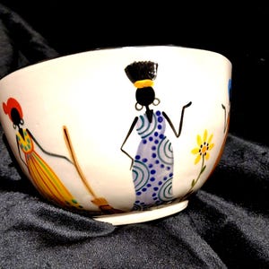 May include: A white ceramic bowl featuring hand-painted illustrations of figures in colourful patterned dresses. One figure wears a yellow dress with a broom, another wears a blue patterned dress, and a yellow flower is painted between them.