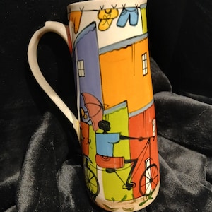 May include: A tall ceramic jug featuring a hand-painted scene of a colourful street with orange, red, and green buildings. The design includes a person riding a bicycle, an umbrella, and laundry hanging on a line above against a cream background.
