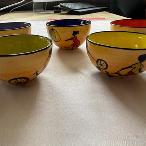 May include: A set of five ceramic bowls with hand-painted designs. The bowls feature vibrant yellow, orange, and lime green exteriors with black line art illustrations of figures and wheels. Each bowl has a contrasting solid-coloured interior.