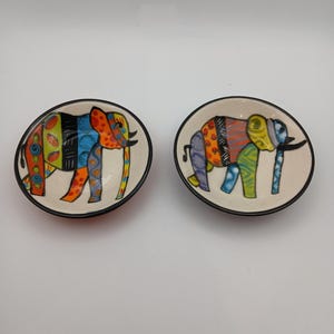 May include: Two round ceramic bowls featuring hand-painted elephant illustrations. Each elephant is decorated with vibrant, multi-coloured geometric patterns, including stripes, dots, and swirls in shades of orange, blue, green, and yellow.