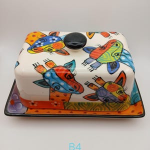 May include: A rectangular ceramic butter dish with a lid featuring a colourful giraffe pattern in shades of orange, blue, yellow, and green. The lid has a black knob handle, and the base has an orange border with black trim. The text B4 is visible.