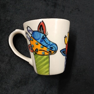 May include: White ceramic coffee mug with colourful giraffe illustrations. The giraffe heads are painted with blue, orange, and yellow spots, with green and yellow stripes at the base. The mug has a white handle and a black rim.