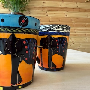 May include: Two ceramic mugs with elephant designs. The mugs feature a black elephant silhouette on an orange background, with blue and yellow accents. One mug has a handle. The mugs are on a white surface.