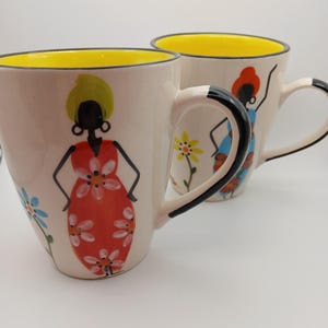 May include: Two off-white ceramic mugs with bright yellow interiors and black-rimmed handles. Each mug features a hand-painted figure wearing a floral dress, with one figure in a red dress and the other in a blue dress, surrounded by small flowers.