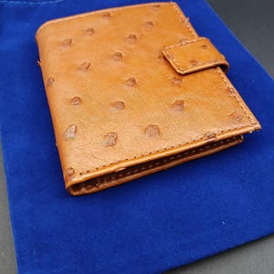 May include: A brown leather wallet with a textured surface and a snap closure. The wallet is on a blue velvet pouch. The wallet appears to be new and unused.