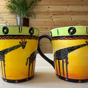 May include: Two ceramic mugs with black handles and a giraffe design. The mugs have a yellow background with a black giraffe illustration, accented by orange and black details. The rims are lime green with black and yellow circular patterns.