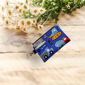 May include: A blue zipped pouch with a construction vehicle pattern. The pouch features images of a bulldozer, cement mixer, and other vehicles. It has a black wrist strap and a silver-coloured clasp.