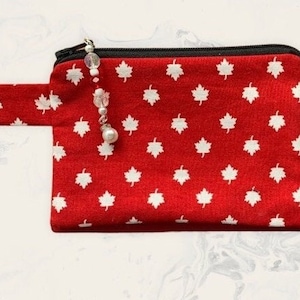 May include: A red zippered pouch with a white maple leaf pattern. The pouch has a black zipper and a small fabric loop on the side. A decorative charm with beads and a pearl hangs from the zipper.