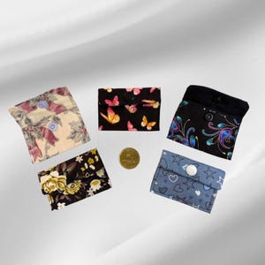 May include: Five small fabric pouches with snap closures, featuring various patterns including butterflies, flowers, stars, hearts, and paisley designs, arranged around a single gold-coloured coin for scale, showcasing diverse textile prints and colours.