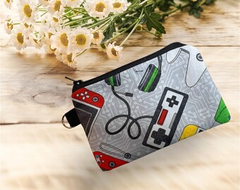 May include: A gray zippered pouch with a black zipper and a black loop. The pouch is patterned with video game controllers, headphones, and circuit board designs in red, green, yellow, and white. The background is a light wood surface.